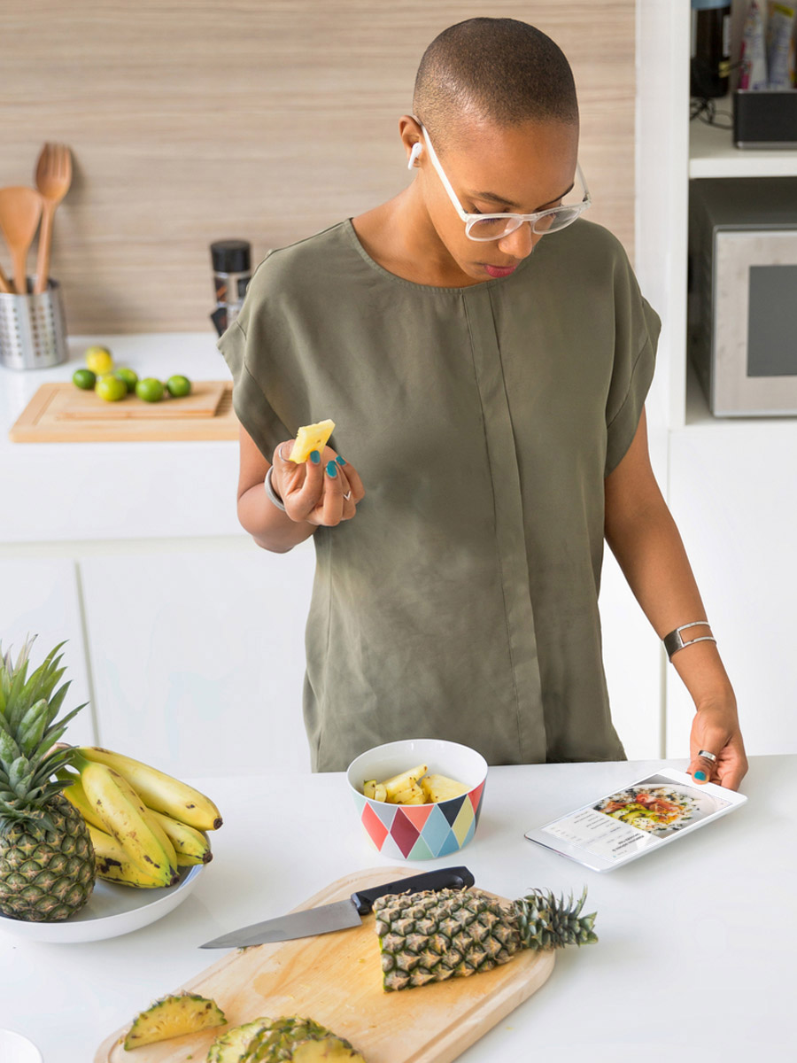 young modern woman standing at kitchen counter referring to foodrush app on her tablet while snacking on pineapple